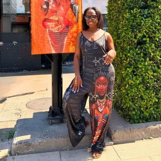A woman wearing a sleeveless wide-leg jumpsuit with a bold graphic print of a Black woman's face and geometric patterns, standing on a city sidewalk next to a green hedge and an orange poster.