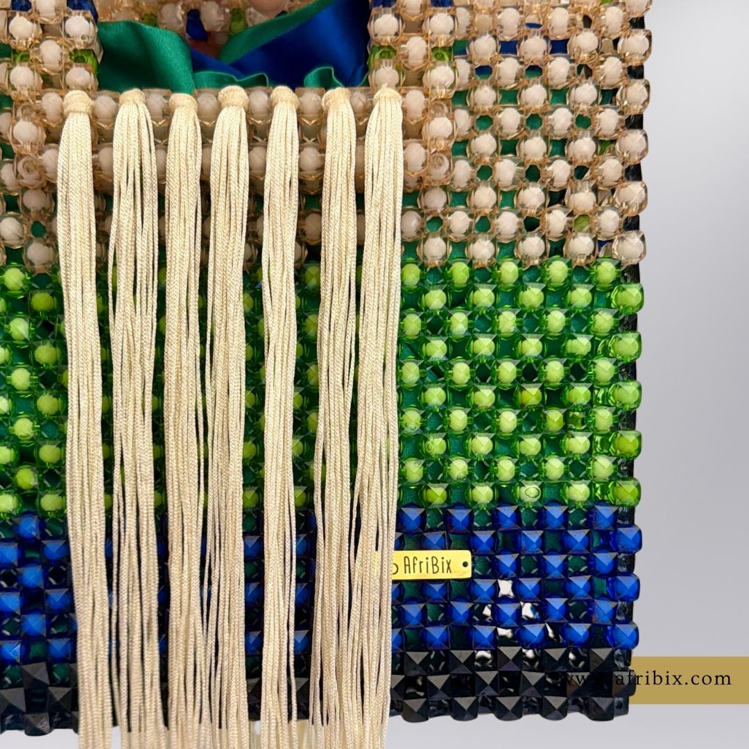 A close-up of a handcrafted beaded purse or bag with cream tassel fringe, featuring horizontal bands of cream beads, green faceted beads, and blue and black beads, with a small yellow tag reading 'AfriBix'.