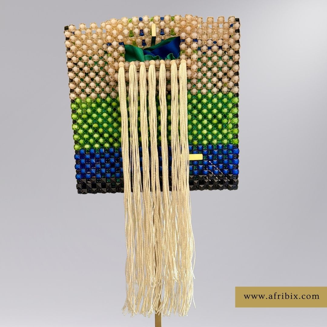 A square beaded clutch bag with a geometric pattern of beige, green, blue, and black beads, featuring a long cream-colored fringe tassel hanging from the center. The clutch is displayed on a stand against a neutral gray background.