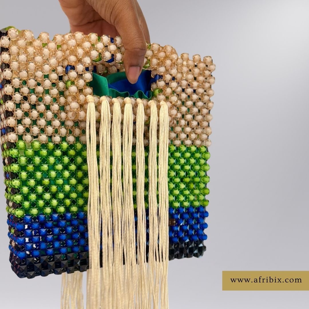 A hand holds a handmade beaded clutch bag with a geometric striped pattern in beige, green, blue, and black, featuring long cream-colored fringe tassels. A website watermark is visible in the bottom right corner.